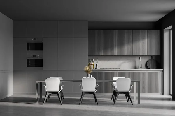 Modern kitchen interior with gray walls, a concrete floor and gray and wooden countertops. A long table with chairs near it. 3d rendering