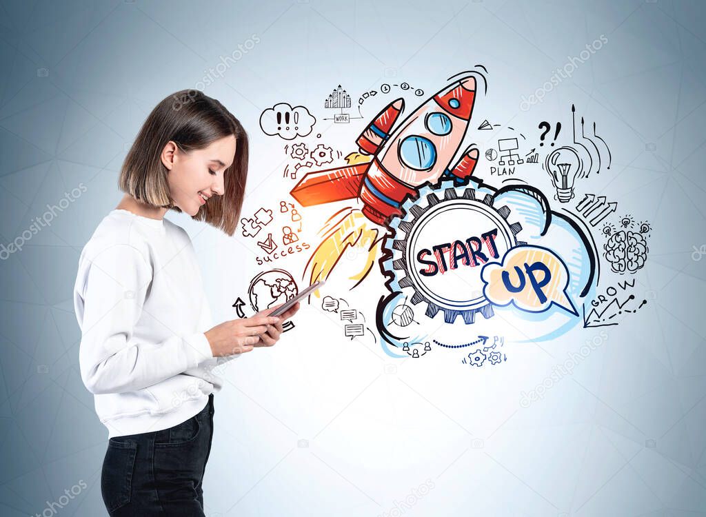 Side view of young woman using tablet computer near gray wall with colorful start up sketch drawn on it. Concept of success and new business project launch