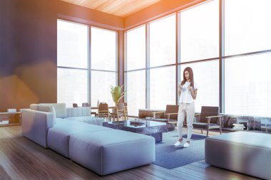 Kahve içen ve sosyal ağları kontrol eden bir kadın, oturma odasının modern panoramik lüks içinde Smartphoneon kullanıyor. Güneş ışınları mercekleri odayı dolduruyor..