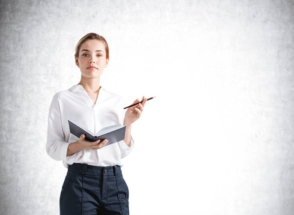 Портрет привлекательной деловой женщины в формальной одежде, стоящей перед бетонной стеной, держащей блокнот и интересующейся мерами по улучшению бизнеса.