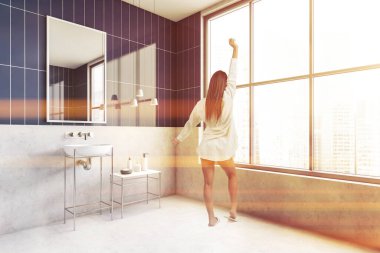 Büyük beden gömlek giyen esmer kız kameranın karşısında duruyor modern banyo, uyandıktan sonra ellerini uzatıyor ve panoramik pencereden dışarı bakıyor.