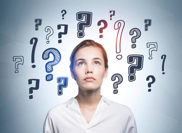 Европейская привлекательная молодая женщина в белой рубашке смотрит вверх и размышляет о карьерных возможностях. На стене сзади нарисованы вопросительные знаки. Концепция поиска ответа.