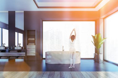 Beyaz küvetli siyah ve ahşap oda ve aynalı lavabo, köşede bitki. Döşemeli döşeme ve pencerenin yanında duran kadın, parlayan mercek, renkli görüntü