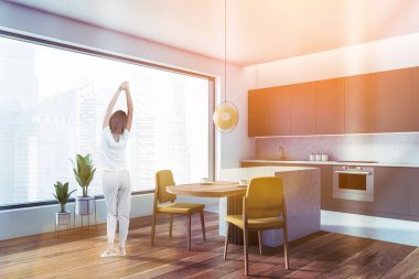 Sabahları pencerenin yanında genç bir kadın mutfak takımlı bir yemek odası panoramik pencere, yan görüş. Sarı sandalyeli masa, gökdelenlerin manzarası.