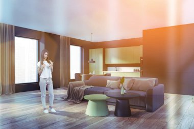 Genç bir kadın telefon kullanıyor, evin içinde kahve içiyor. Parke zeminde gri bir kanepe, yan görüş. Mutfak rafı, yemek masası ve gökdelenlerin şehir manzarası.