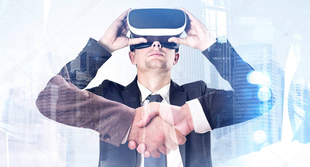 Young handsome businessman in suit holding VR glasses. Double exposure, skyscrapers and digital handshake as a concept of enter into business agreement. Concept of corporate network and virtual reality.