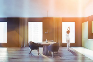 Sabahları pencerenin yanında, yemek odasının içinde mutfak seti olan bir kadın. Üç gri sandalyeli ahşap masa, gökdelenler şehir manzaralı.