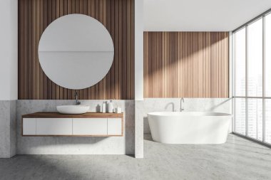 Lavabosu ve beyaz küveti olan banyo, beton zemin ve panoramik pencereli ahşap duvar. Yuvarlak ayna, jel ve sabun ile modern stil oda iç mimarisi.