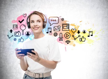 Ellerinde akıllı telefonuyla kulaklıkla gülümseyen günlük kıyafetli bir öğrenci. Sosyal iletişim simgeleri, multimedya, teknoloji ve arayüz kavramı