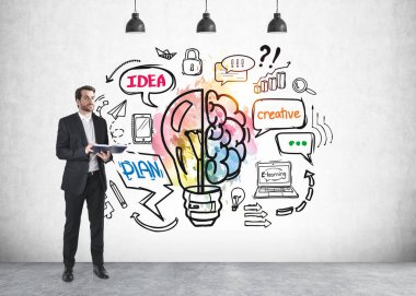 Ofisteki adam elinde defter, parlak fikirler ve renkli beyinle ampul, ofis odasındaki beton duvara çizilmiş planlar ve mesajlar tutuyor. Fikir ve plan kavramı