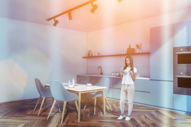 Modern mutfak odasında telefonu olan, yemek masası, gri sandalyeli, lens takan çekici genç bir kadın. Lavabo ve soba, mutfak gereçleriyle dolu dolap.