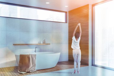 Lüks banyo, banyo küveti ve şehir manzaralı panoramik pencere. Pencerenin yanında duran rahat giysili kadın uyandı. Lens parlıyor, renkli bir görüntü.