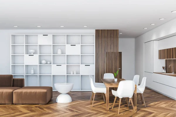Stylish dining room interior with white and wooden walls, wooden floor, long table with white chairs and white counters with built in appliances. Brown sofa and bookcase. 3d rendering