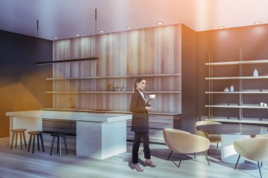 Attractive businesswoman in formal suit is drinking coffee at corporate office kitchen. Modern kitchen set and dining area as facilities for employees. Sun rays filling the room