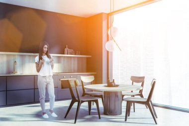 Kadın internete telefon kullanarak ve modern mutfak odasında panoramik pencereli, şehir merkezindeki gökdelenler şehir manzaralı, mercekli kahve içerek bakıyor. Sandalyeler ve yuvarlak masa