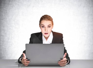 Resmi giysiler içinde, masanın önünde dizüstü bilgisayarın önünde oturan hayrete düşmüş genç iş kadınının portresi. Şirket yaşam tarzı kavramı. Beton duvar.