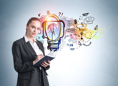 Resmi takım elbiseli, kendine güvenen çekici bir iş kadını elinde bir planlamacı tutuyor ve not alıyor. Yakındaki açık mavi duvarda renkli bir iş fikri olan ampul eskiziyle yeni bir başlangıç yapmayı düşünüyorum. Beyin fırtınası.