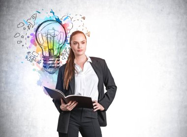 Resmi takım elbiseli, kendine güvenen çekici bir iş kadını elinde bir planlayıcıyla renkli bir iş fikri ampulü çizimiyle yakınlardaki beton duvarda yeni bir başlangıç hakkında kafa patlatıyor. Beyin fırtınası konsepti.