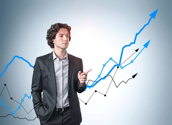 Confident Hispanic businessman in suit pointing out his finger and pondering about business growth in sales of the company. Growing line charts on light blue wall