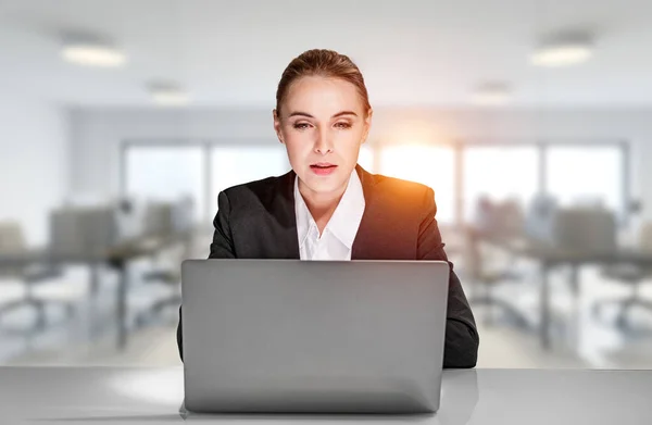 Attractive business woman is sitting at workplace in front of the laptop, leaning on the desk and wondering about analyzing business report. Concept of working from panoramic office
