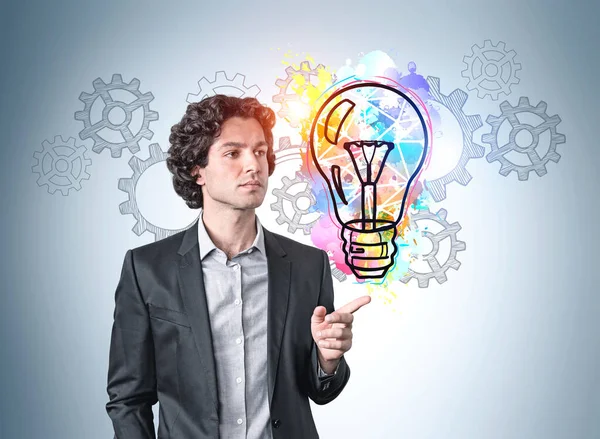 Hispanic handsome businessman in suit pointing out by finger and pondering about new ideas. Blue wall with colorful business idea light bulb and cog wheels sketches. Creativity and brainstorming