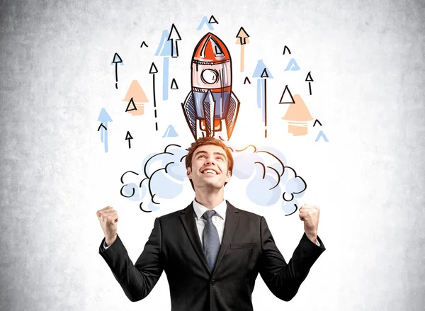 Portrait of a happy young man in business suit with both hands up and dreaming about launch a business start up. Concept of success. Colorful rocket sketch drawing on wall