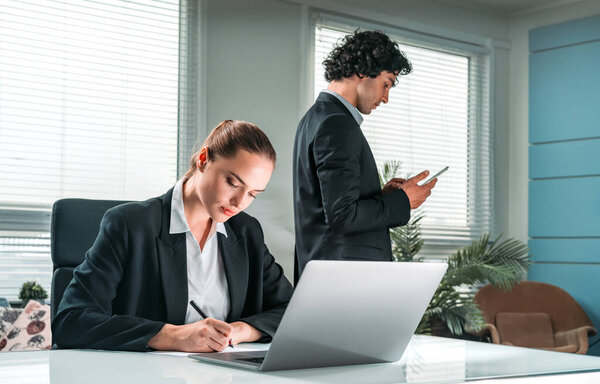 Two working concentrated business people, one is sitting at the desk in front of laptop, second is using smartphone at office background. Concept of teamwork