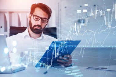 Beyaz gömlekli bir iş adamı panoya bakıyor ve kahve içiyor. Bilgisayarlı ve arka planında akıllı telefon olan bir ofis işyeri. Forex şamdanlar, grafik ve grafik ön planda