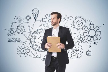 Resmi takım elbiseli bir işadamı elinde bir karalama tahtası ile beyaz çarklı, dişli, ampullü ve el sıkışma ikonlu bir karalama tahtası tutuyor. Yaratıcı iş fikri için düşünme kavramı