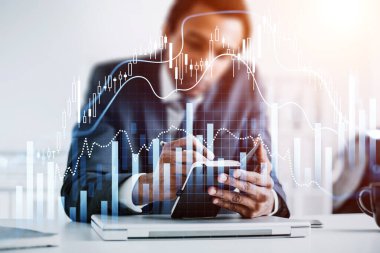Afrikalı Amerikalı iş adamı elinde not defteri tutuyor ve not alıyor. Arkasında dizüstü bilgisayar olan bir ofis işyeri. Ön planda eski şamdan ve finansal grafik var. Ticaret kavramı