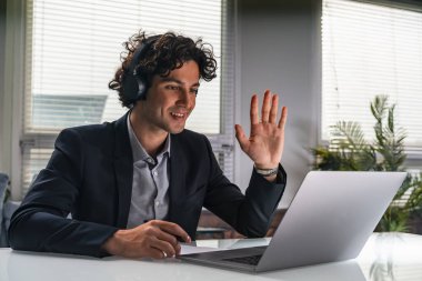 Resmi takım elbiseli gülümseyen kıvırcık iş adamı, video konferans görüşmesi ve meslektaşlarını selamlama yoluyla konuşuyor. Arkasında dizüstü bilgisayar olan bir ofis işyeri. Takım çalışması ve mesafe toplantısı kavramı