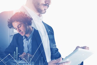 İki işadamı birlikte çalışıp not alıyor ve tablet üzerinde yazıyor. Arka planda ofis işyeri var. Finansal grafik ve çizgi ön planda. Takım çalışması, iletişim ve işbirliği kavramı