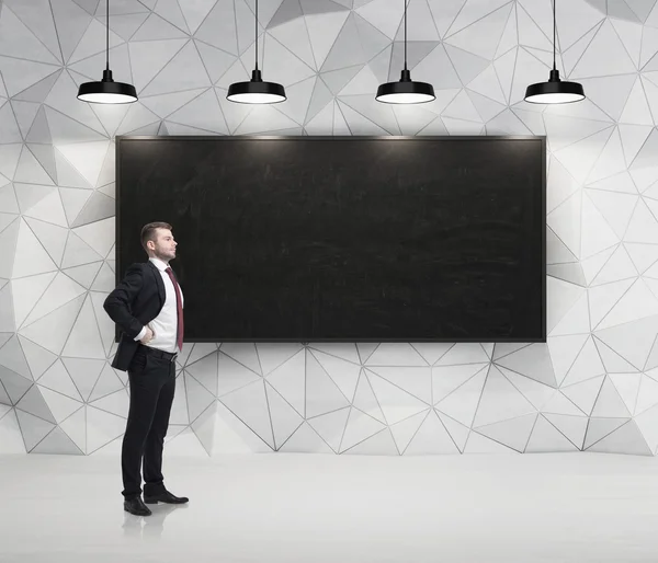 Young successful businessman is standing in front of the chalkboard in the concrete room with four black lamps. — Stock Photo, Image