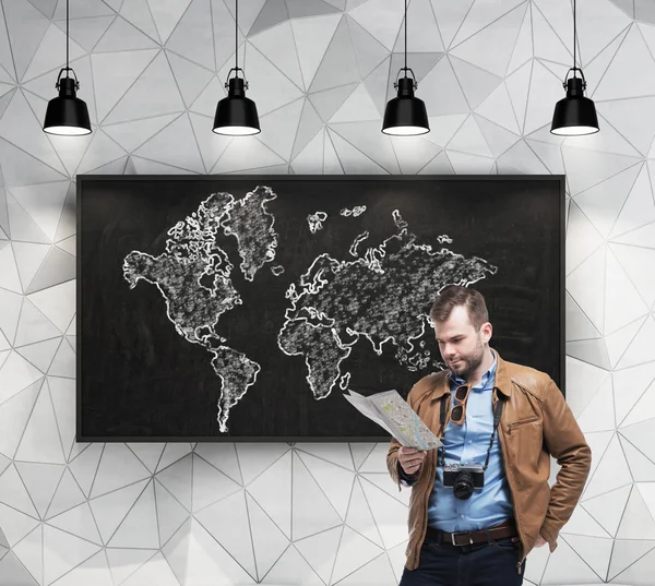 Turista bonito está examinando o mapa na frente do quadro com o esboço do mapa do mundo — Fotografia de Stock