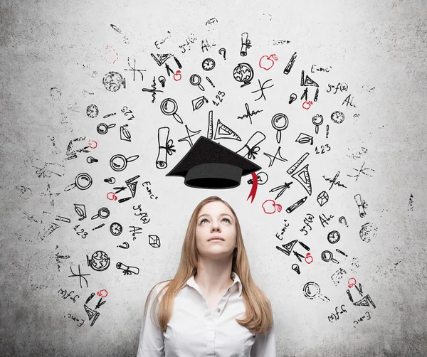 Young beautiful business woman is thinking about education at business school. Drawn business icons over the concrete wall. Graduation hat. — Stock Photo, Image