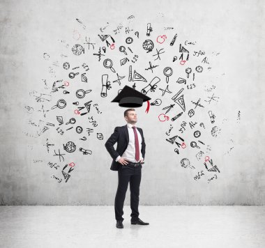 Young handsome businessman is thinking about education at business school. Drawn business icons over the concrete wall. Graduation hat.