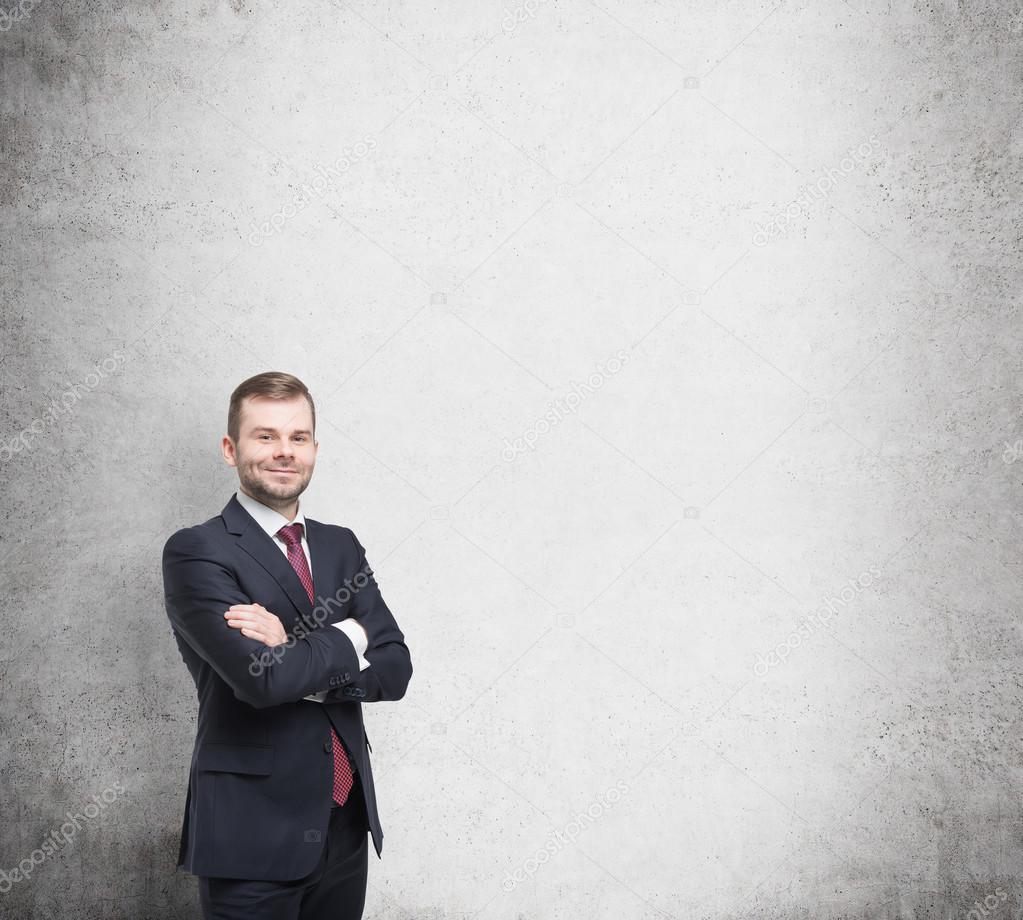 Smiling handsome businessman in a black suit with the fold arms. Concrete wall background.