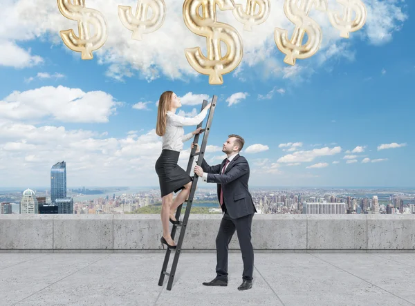 Businessman holds a ladder for the business lady. The concept of the teamwork. On the roof terrace in Manhattan. Dollar icons falling from the sky. — Stock Photo, Image