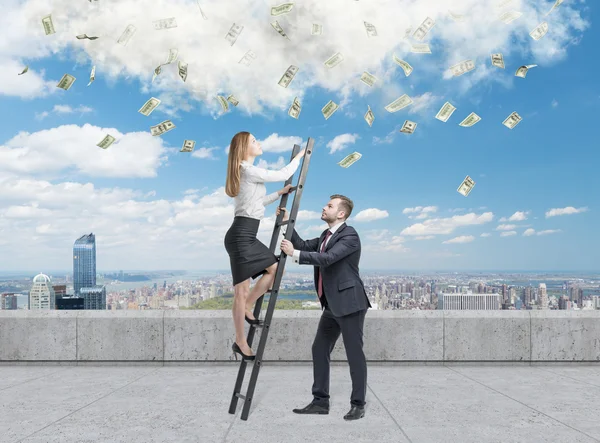 Businessman holds a ladder for the business lady. The concept of the teamwork. On the roof terrace in Manhattan. Dollar notes falling from the sky. — Stock Photo, Image