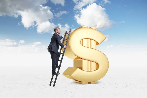 Бизнесмен залезает на огромный золотой знак доллара. Голубое небо на фоне облаков . — стоковое фото