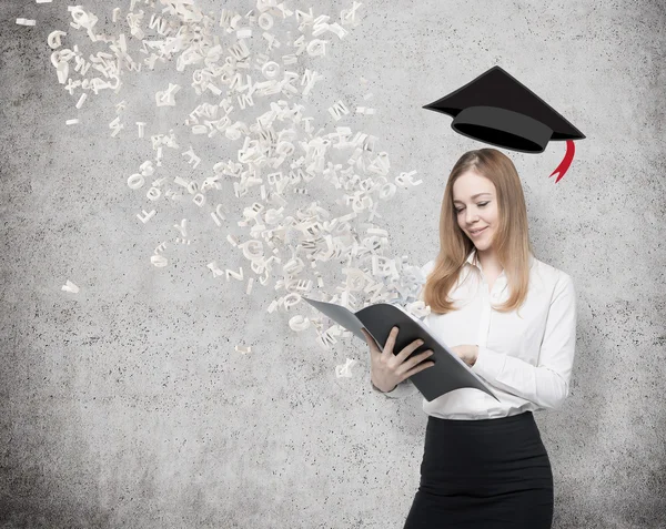 Um estudante bonito está segurando uma pasta preta com letras voando para fora da pasta. Chapéu de formatura por cima da cabeça da senhora. Concreto fundo . — Fotografia de Stock