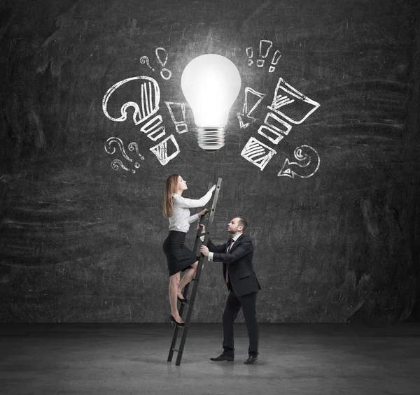 A handsome manager assists a beautiful colleague to climb up the stairs to reach new business ideas. A light bulb as a concept of new ideas. — Stock Photo, Image