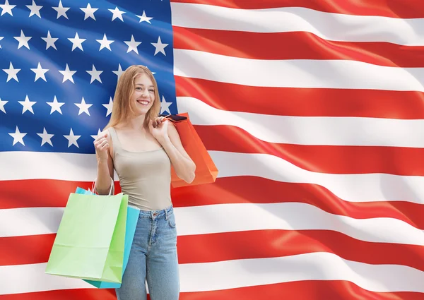 En glad ung kvinna med de färgglada påsarna från fancy butiker. oss flagga bakgrund. — Stockfoto