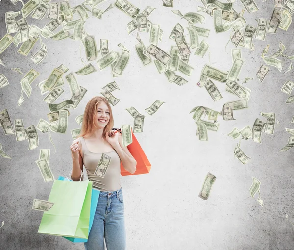 Eine fröhliche junge Frau mit den bunten Einkaufstüten aus den schicken Geschäften. Dollarnoten fallen von der Decke. Konkreter Hintergrund. — Stockfoto