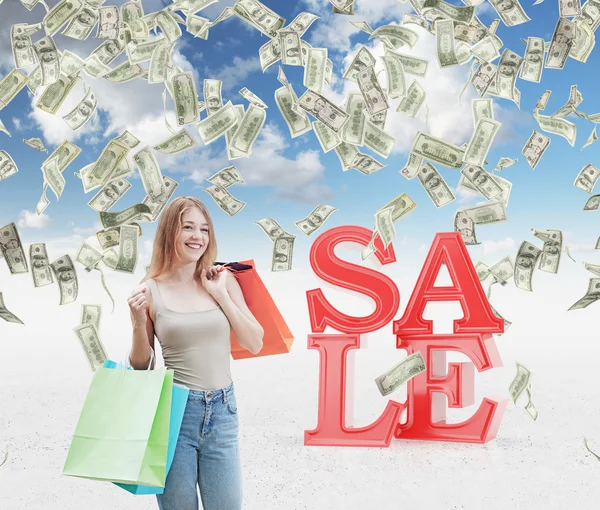 फॅन्सी शॉपमधून रंगीबेरंगी शॉपिंग पिशव्या असलेली एक आनंदी तरुण स्त्री. विक्री आणि सूट ही संकल्पना म्हणून डॉलरच्या नोटा आकाशातून खाली येत आहेत. ठोस पार्श्वभूमी . — स्टॉक फोटो, इमेज