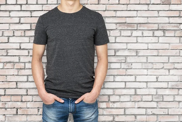 Man wearing dark grey t-shirt, brick wallbackground. Hands in the pockets.