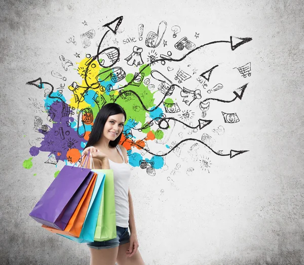 Beautiful smiling young woman with the colourful shopping bags from the fancy shops. Concrete background with drawn shopping icons. — Stock Photo, Image