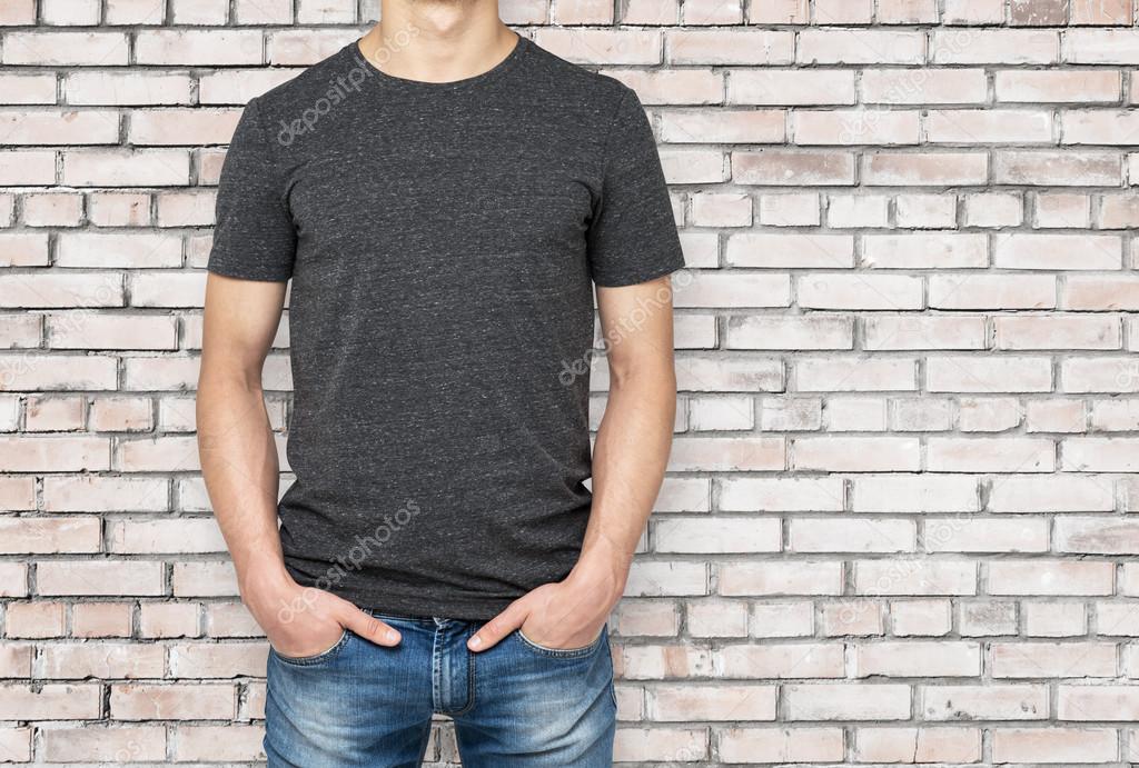 Man wearing dark grey t-shirt, brick wallbackground. Hands in the pockets.