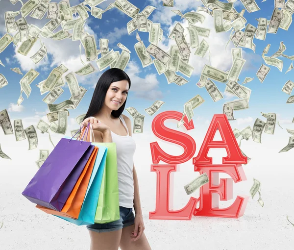 Beautiful smiling brunette woman with the colourful shopping bags from the fancy shops. Falling dollar notes from the cloudy sky. A concept of discounts and sales. — Stockfoto