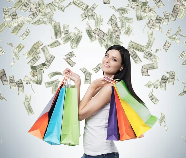 Mulher morena sorridente bonita com os sacos de compras coloridos das lojas extravagantes. Queda de notas de dólar do teto . — Fotografia de Stock